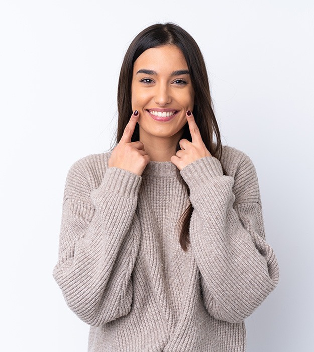 Woman in brown sweater pointing to her smile with both hands